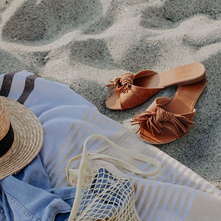 Sandals laying in the sand next to a bag, sun hat and blanket